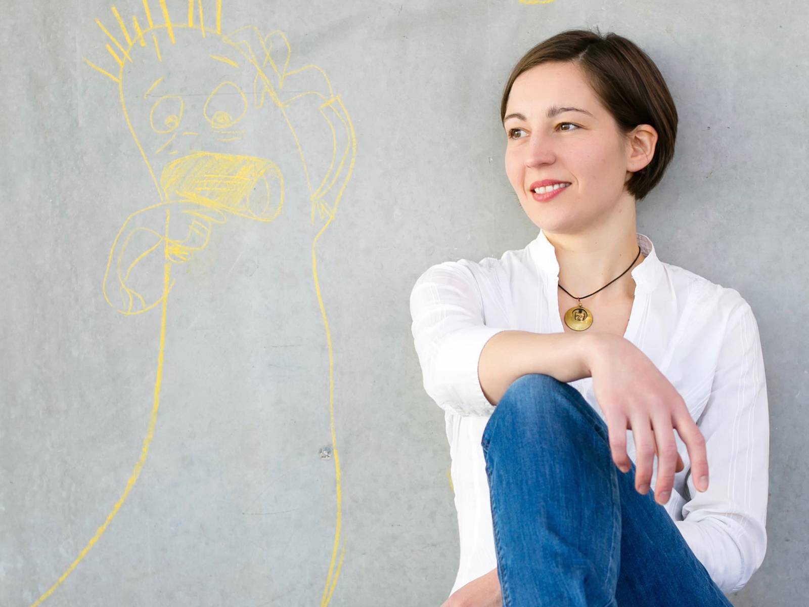 Portraitaufnahme einer Frau, die vor einer grauen Wand sitzt, neben ihr ist mit gelber Kreide eine Figur aufgemalt